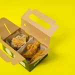 Kotak kardus kemasan makanan terbuka dilihat dari atas dengan latar kuning cerah, ilustrasi box snack kecil untuk usaha kuliner